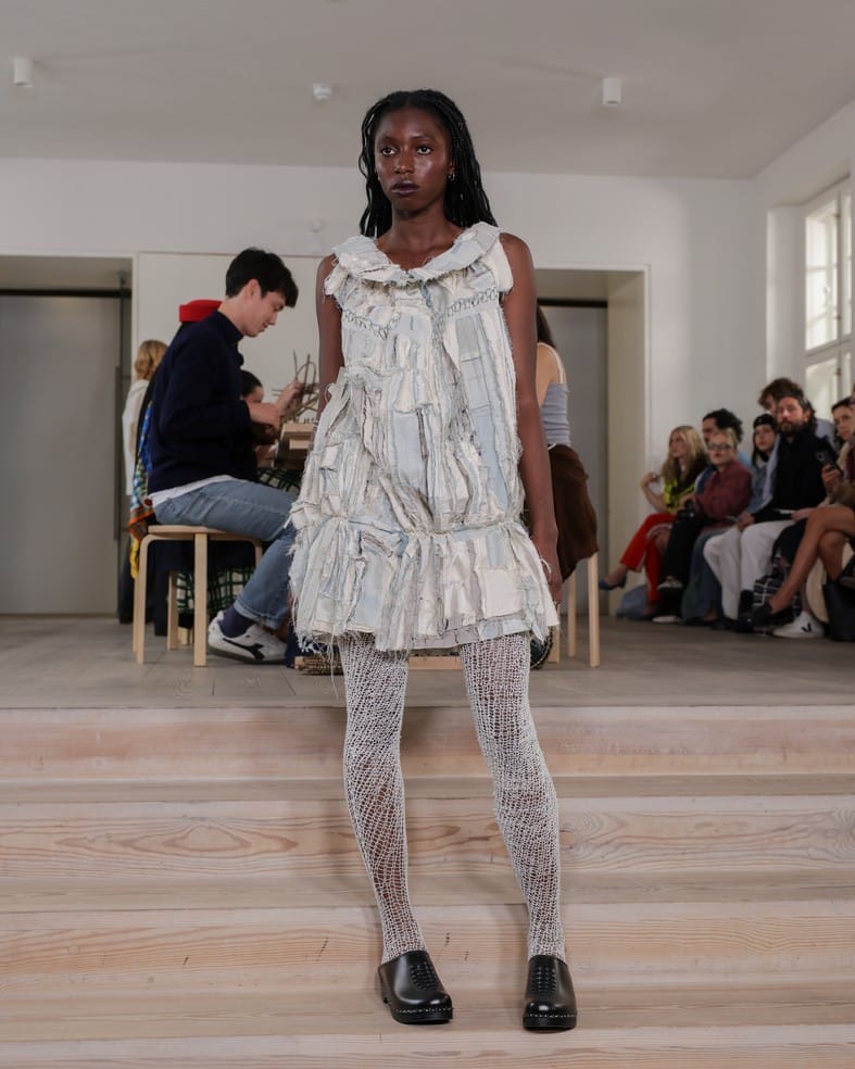 A model with long braided hair wears a textured, layered white dress and patterned white tights with black shoes, walking indoors on wooden steps. Several seated people watch in the background.