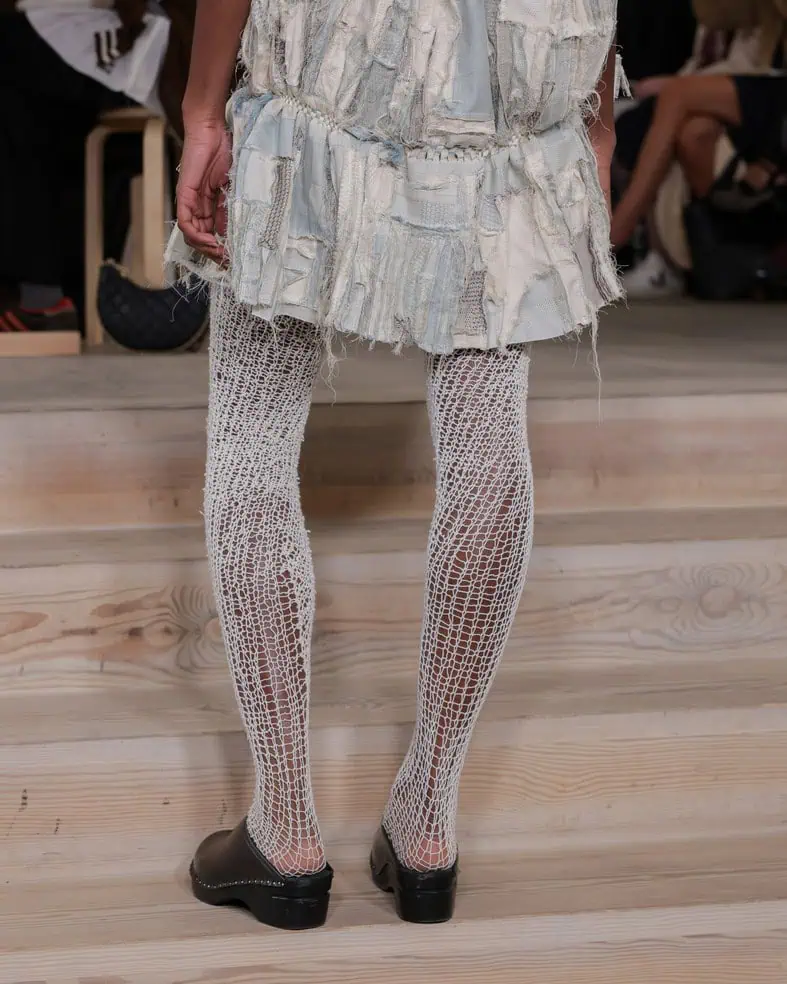 A person wearing a patchwork skirt, white knit tights, and black clogs stands on wooden steps. The focus is on their legs and outfit details; the background shows people seated.