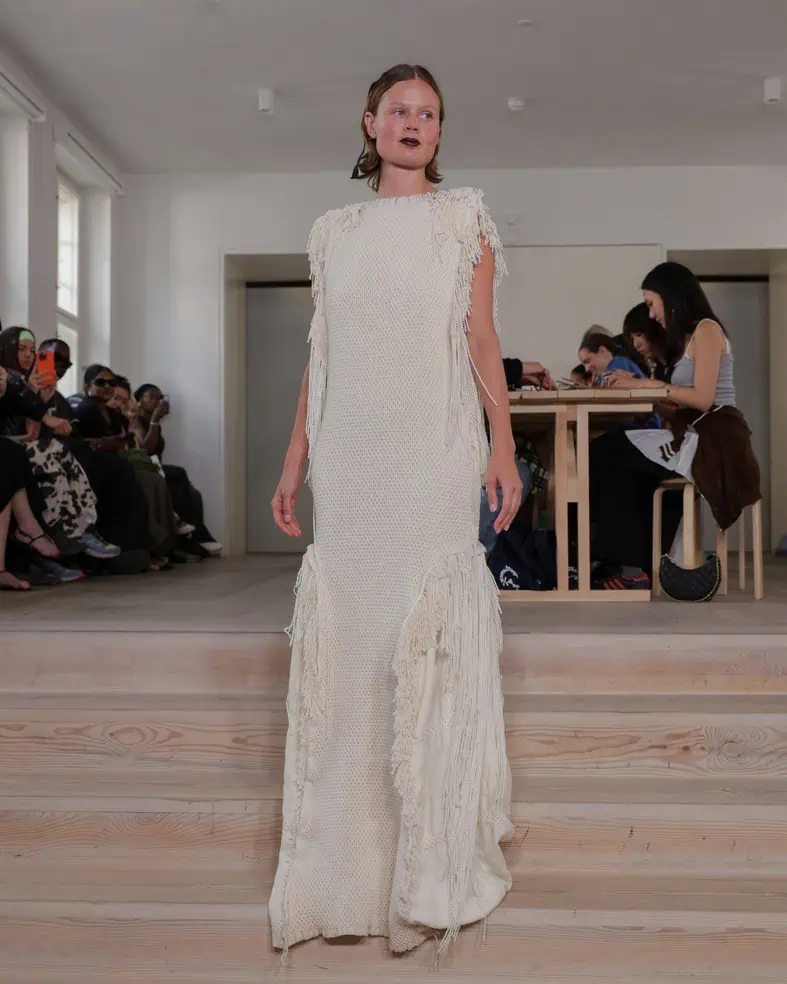 A model walks indoors wearing a long, off-white textured gown with fringed details, while seated people observe the fashion show in the background.