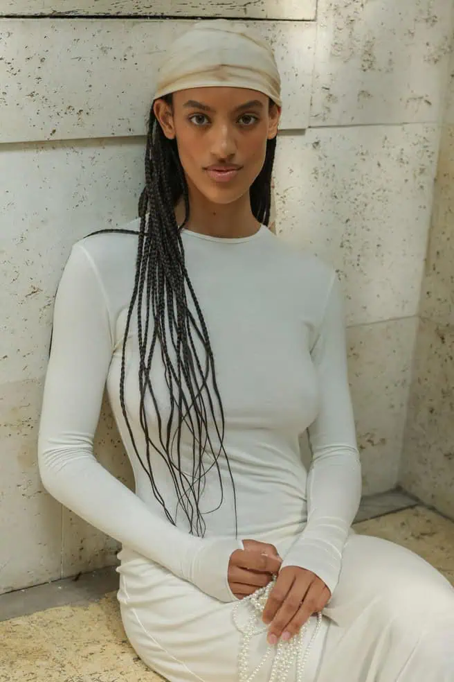 A woman with long braided hair, wearing a cream-colored headscarf and a fitted long-sleeve white dress, is seated against a textured stone wall, holding a beaded Taus necklace in her lap.