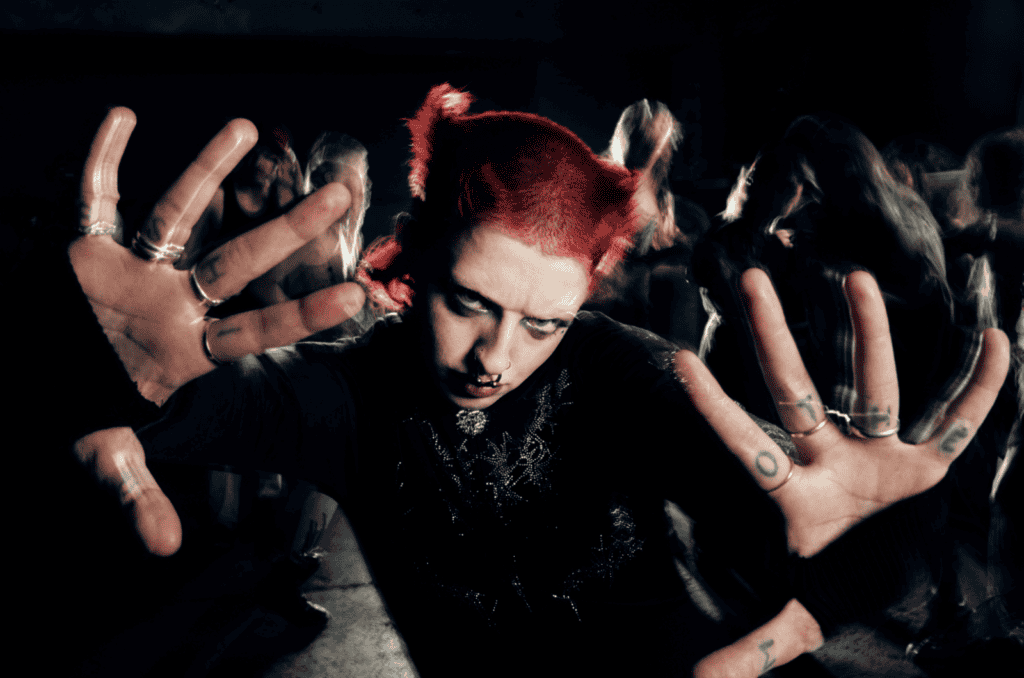 Cody Frost with bright red hair, dark eye makeup, facial piercings, and rings on their fingers poses with hands outstretched toward the camera at a crowded event, with people blurred in the background.
