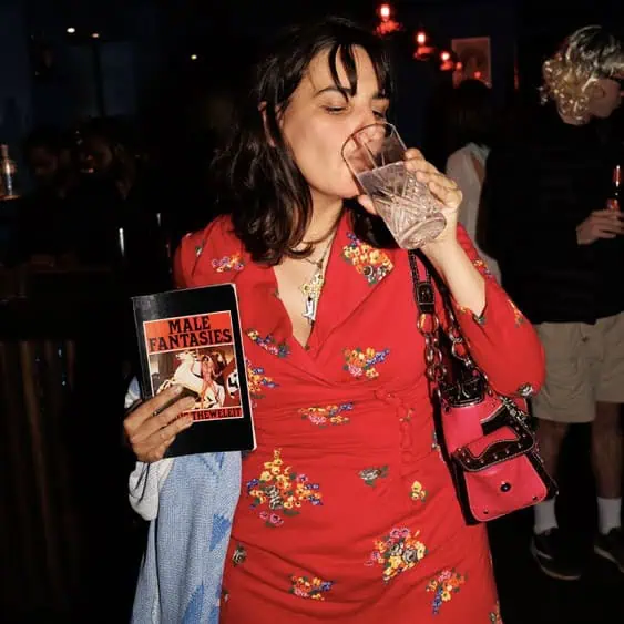 A woman in a red floral dress holds a book titled Male Fantasies and drinks from a glass at a dimly lit bar or party. She has a pink purse over her shoulder and wears a necklace. Other people are in the background.