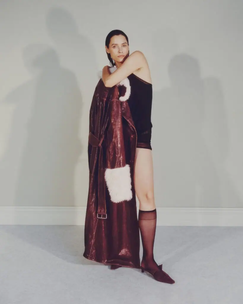 A person stands indoors against a light-colored wall, wearing a black sleeveless top, black shorts, sheer black knee-highs, and brown pointed shoes, while holding a long brown coat with light fur trim draped over one arm.