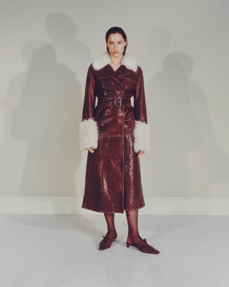 A woman stands against a light-colored wall, wearing a long brown leather coat with a wide belt, white faux fur collar and cuffs, black tights, and brown heeled shoes. Her expression is neutral.