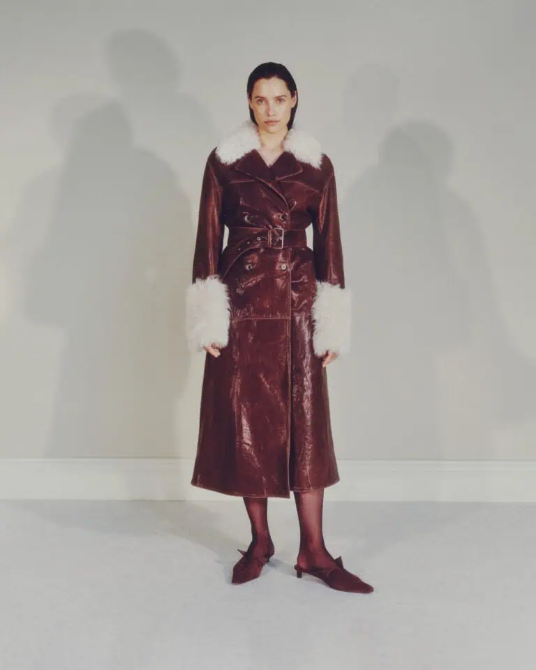 A woman stands against a light-colored wall, wearing a long brown leather coat with a wide belt, white faux fur collar and cuffs, black tights, and brown heeled shoes. Her expression is neutral.