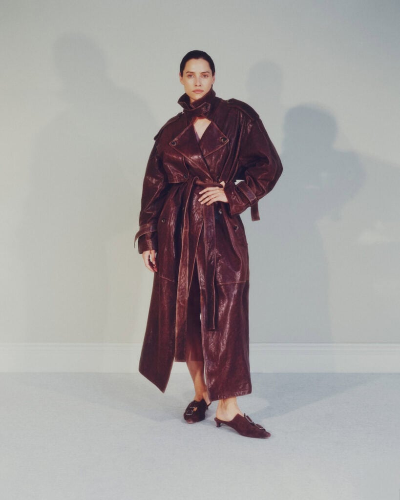 A person stands against a light gray wall wearing a long, dark brown leather trench coat with a belt and heeled shoes. The coat is partially open at the top, and the person looks directly at the camera.
