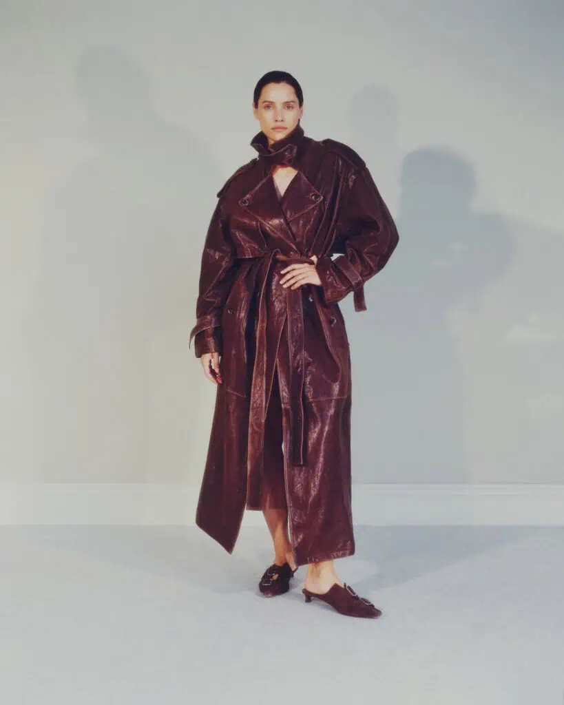 A person stands against a light gray wall wearing a long, dark brown leather trench coat with a belt and heeled shoes. The coat is partially open at the top, and the person looks directly at the camera.