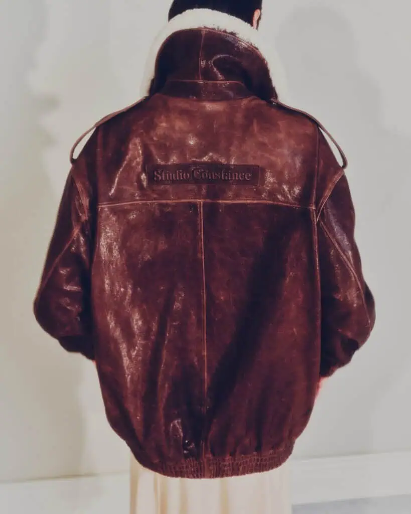 A person with short dark hair, seen from behind, wears a brown leather jacket with Studio Constance embroidered on the back and a white shearling collar. The background is plain and softly lit.