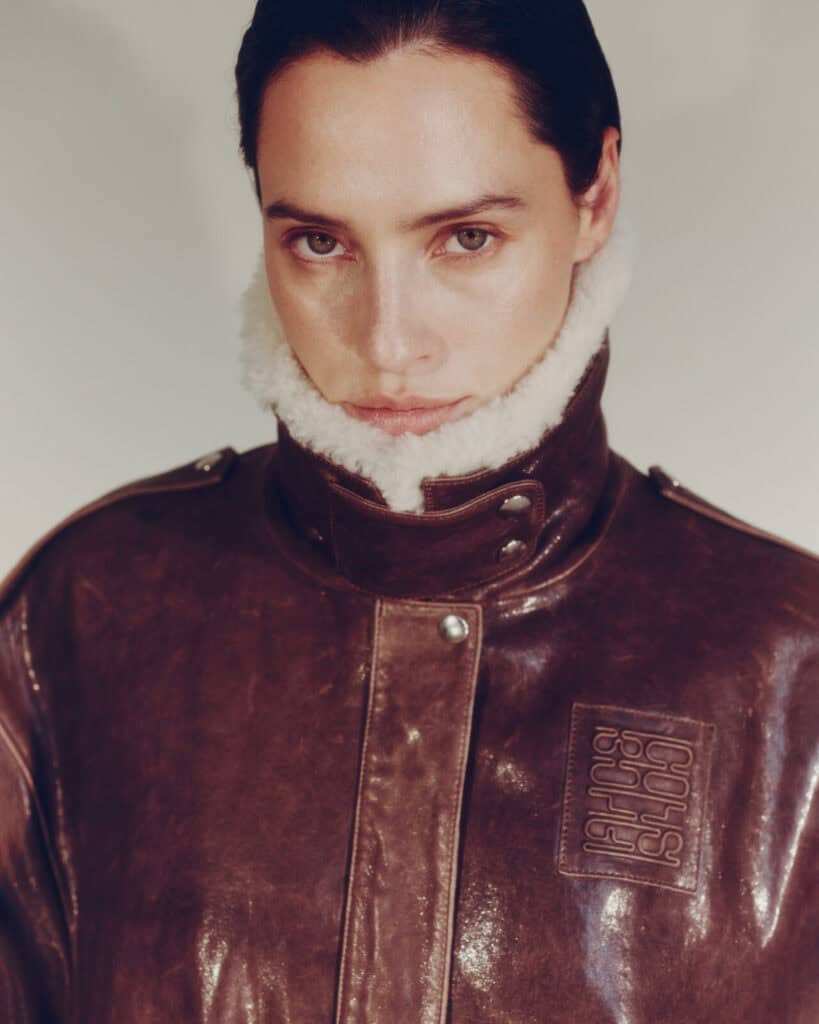 A person with dark hair pulled back wears a brown, shiny leather jacket with a high, white shearling collar. The jacket has a patch with raised lettering, and the person looks directly at the camera with a neutral expression.