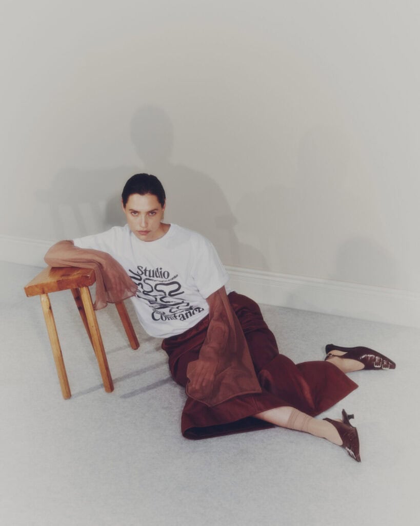 A woman sits on the floor, leaning against a wooden chair. She wears a white graphic T-shirt, a brown skirt, sheer brown tights, and pointed brown heels, with a brown jacket draped over her arm. The background is neutral.