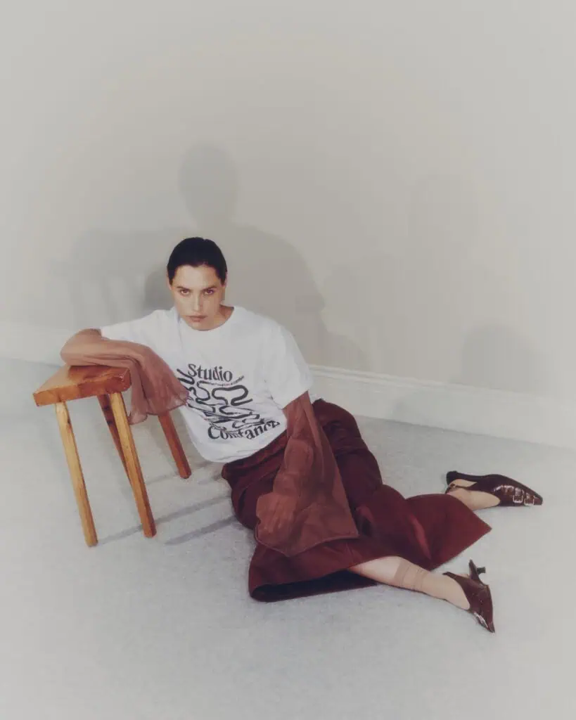 A woman sits on the floor, leaning against a wooden chair. She wears a white graphic T-shirt, a brown skirt, sheer brown tights, and pointed brown heels, with a brown jacket draped over her arm. The background is neutral.