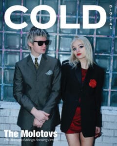 The molotovs stand against a glass brick wall. The man wears sunglasses, a suit, and tie; the woman wears a black blazer with a red rose pin over a red dress. The magazine cover reads: COLD DIGITAL and The Molotovs.