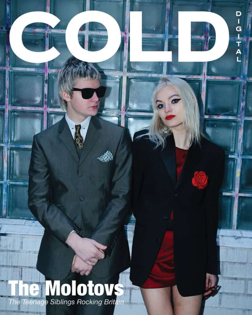 The molotovs stand against a glass brick wall. The man wears sunglasses, a suit, and tie; the woman wears a black blazer with a red rose pin over a red dress. The magazine cover reads: COLD DIGITAL and The Molotovs.