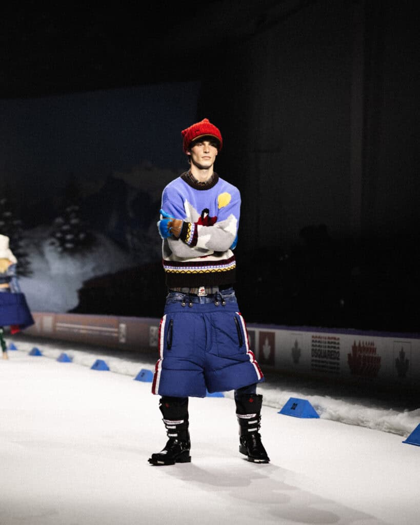 A model wearing a colorful sweater, blue puffy pants, a red knit hat, and black boots walks on a runway resembling a snowy scene, with pine trees and mountains painted in the background.