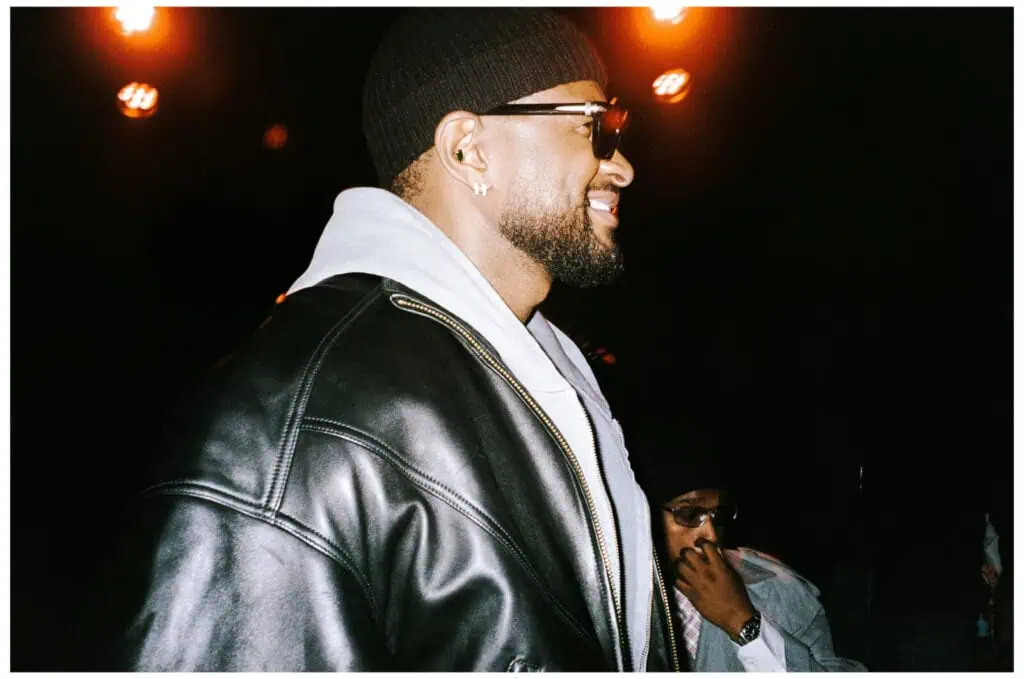 A man in a black beanie, sunglasses, and a leather jacket smiles while standing under orange lights. Another person sits in the background, partially obscured, wearing glasses and a dark hat.