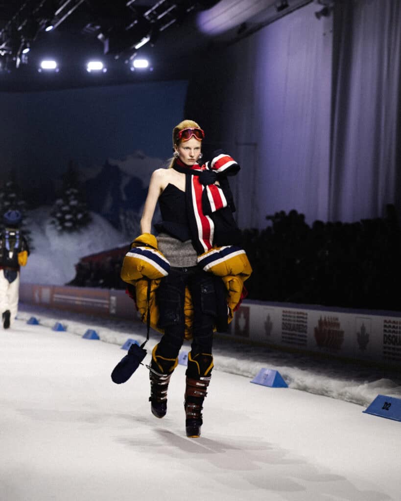 A model walks down a snowy-themed runway wearing bulky, layered winter clothing, including a large yellow puffer jacket draped over one shoulder, black pants, boots, and ski goggles on their head.