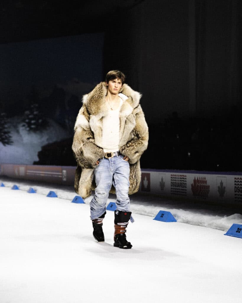 A male model walks a runway wearing a large, fur coat over a white shirt, blue jeans, and black boots. The scene is winter-themed, with snow, trees, and ski slope markers in the background.