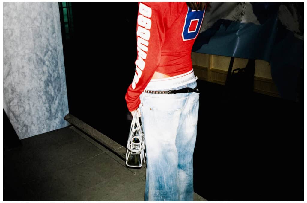 A person wearing a red long-sleeve shirt with white lettering and baggy light-wash jeans stands indoors, holding a white handbag by their side. The scene is dimly lit with marble and dark surfaces in the background.