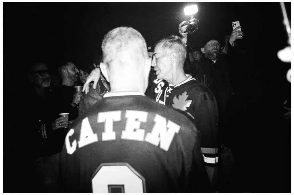 A black-and-white photo of a crowd at night. In the foreground, a person wearing a jersey with CATEN on the back faces away from the camera. People around them laugh, talk, and take photos. One person holds up a phone with flash.