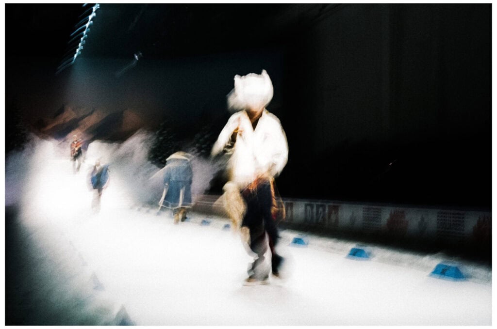 A blurred photo of models walking down a runway, wearing winter clothing and hats; the background suggests a snowy, mountain-like setting. The motion blur creates a dreamlike, dynamic effect.