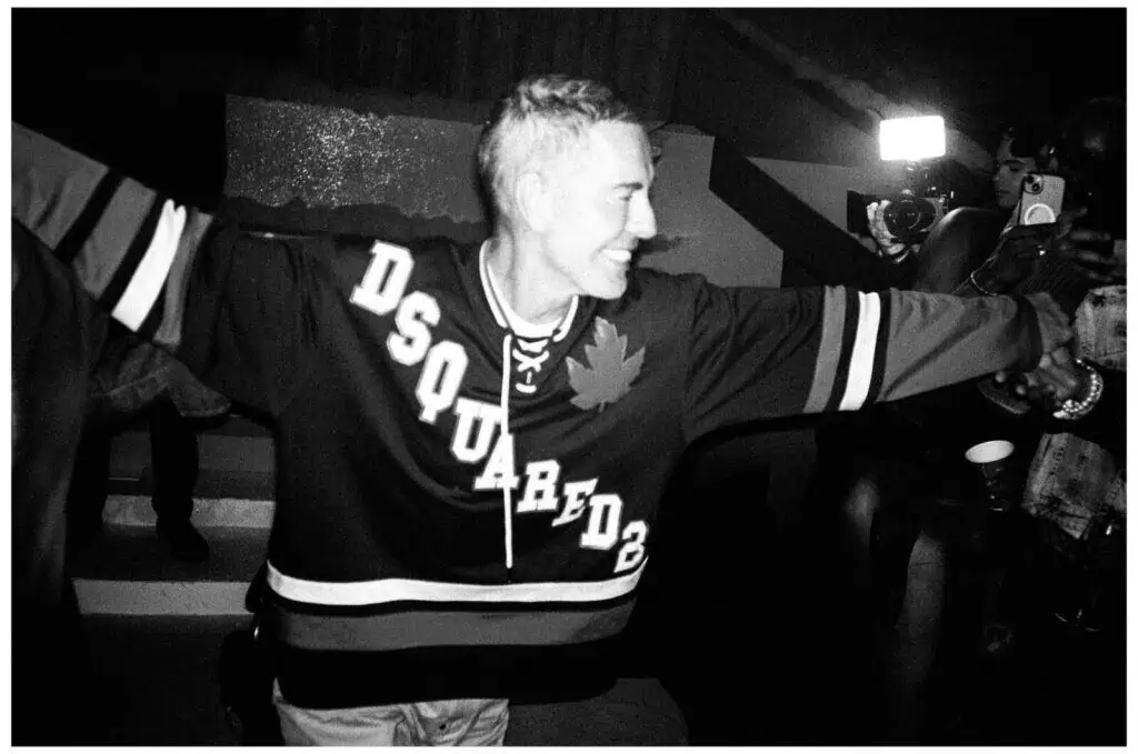 A person wearing a “DSQUARED2” hockey jersey smiles with outstretched arms in a lively, dimly lit setting; photographers and bright flashes are visible in the background.