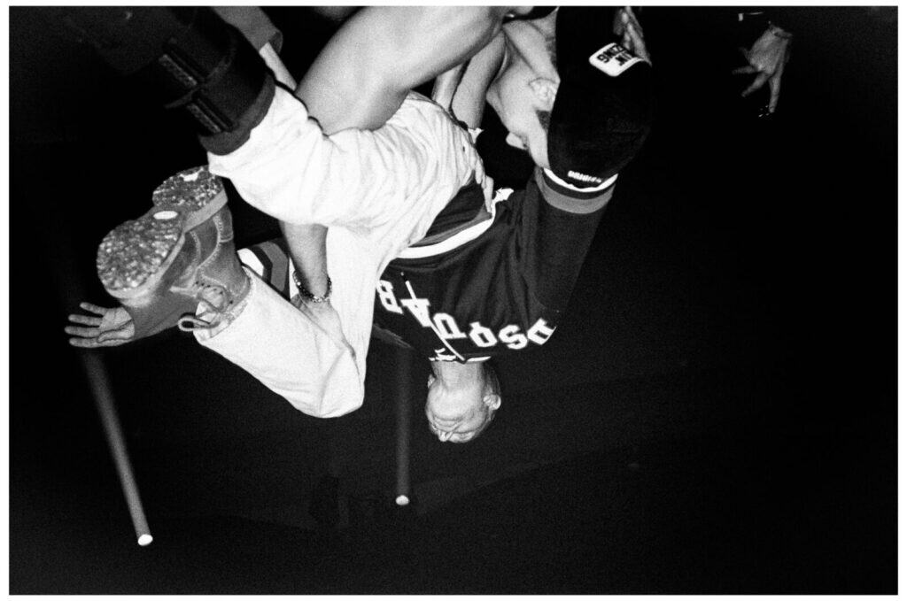 A person wearing boots and a sports jersey is caught mid-air upside down, likely stage diving or crowd surfing at a concert, with outstretched arms and blurred hands visible in the dark background.