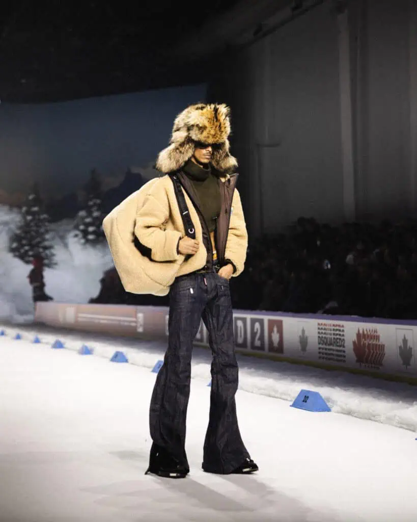 A model walks a winter-themed runway wearing a fur hat, tan shearling jacket, brown top, dark flared jeans, and black shoes, with a large matching tan bag slung over one shoulder. Snowy trees and an audience are in the background.