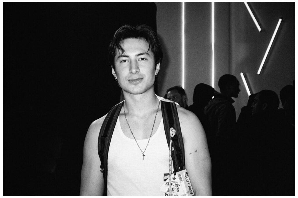 A young man wearing a white tank top and a backpack stands indoors, looking at the camera. Neon lights and silhouettes of people are visible in the background. The image is in black and white.
