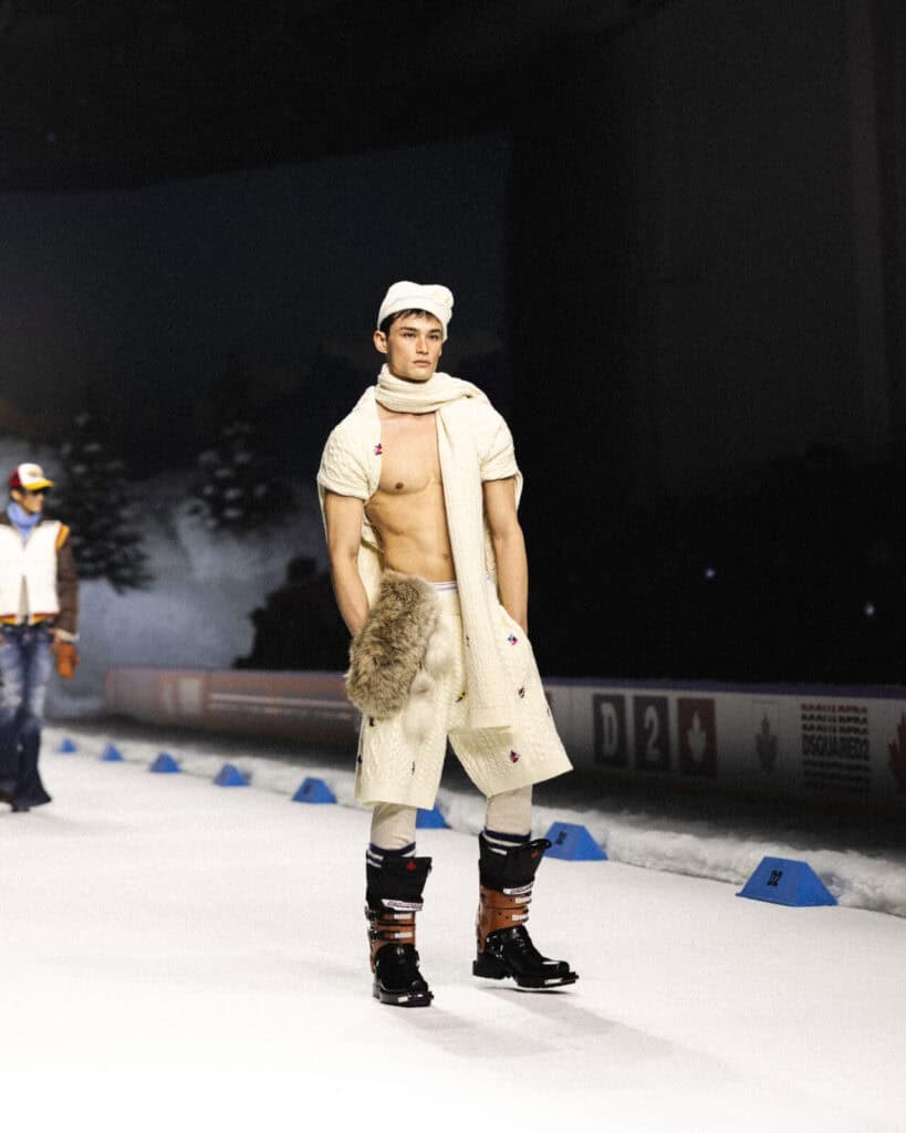 A male model walks on a snowy runway wearing a white knit scarf and cap, cream pants, black boots, and a fur accessory; his chest is bare and trees are visible in the background.
