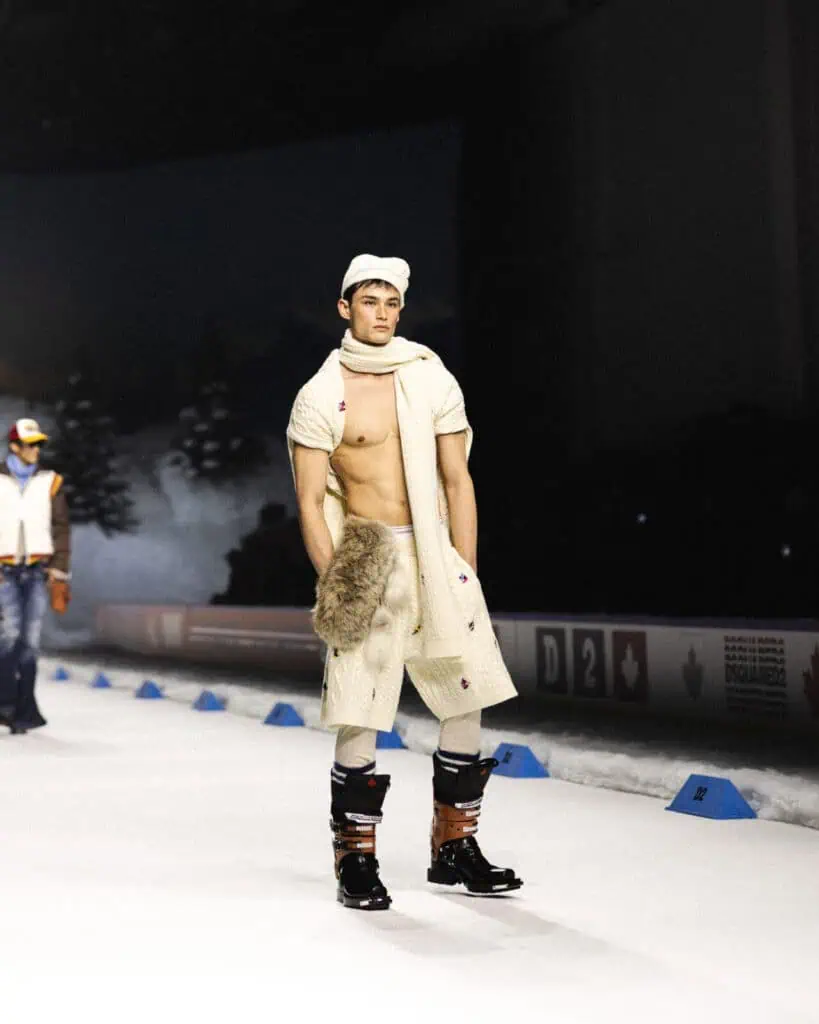A male model walks on a snowy runway wearing a white knit scarf and cap, cream pants, black boots, and a fur accessory; his chest is bare and trees are visible in the background.