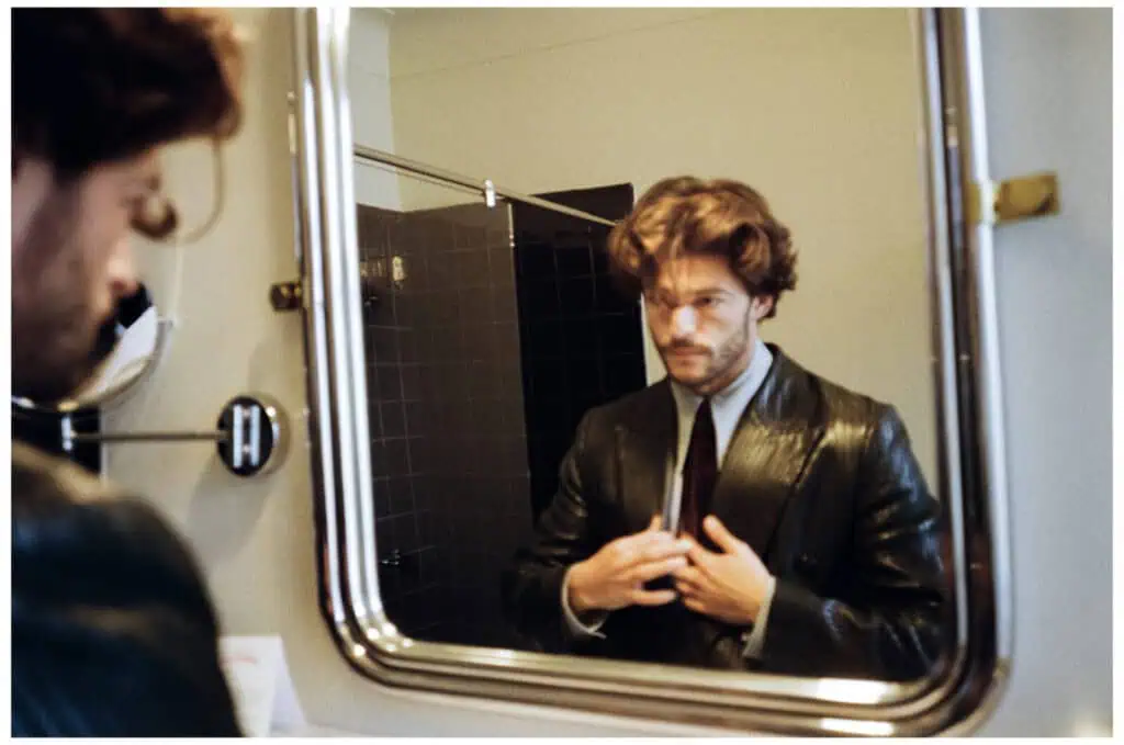 A man in a suit adjusts his jacket while looking at himself in a bathroom mirror. His reflection is visible, and the bathroom features neutral colors and tile walls.