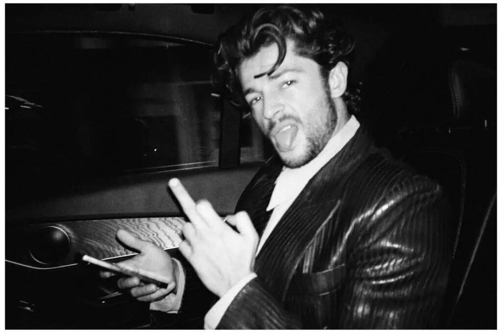A man in a pinstripe suit sits in a car, holding a phone and making a playful face while sticking out his tongue and raising his middle finger toward the camera. The photo is black and white.
