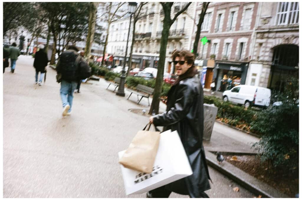 A person in a black leather coat and sunglasses walks down a city sidewalk, holding several shopping bags. Buildings, parked cars, and other pedestrians are visible in the background.