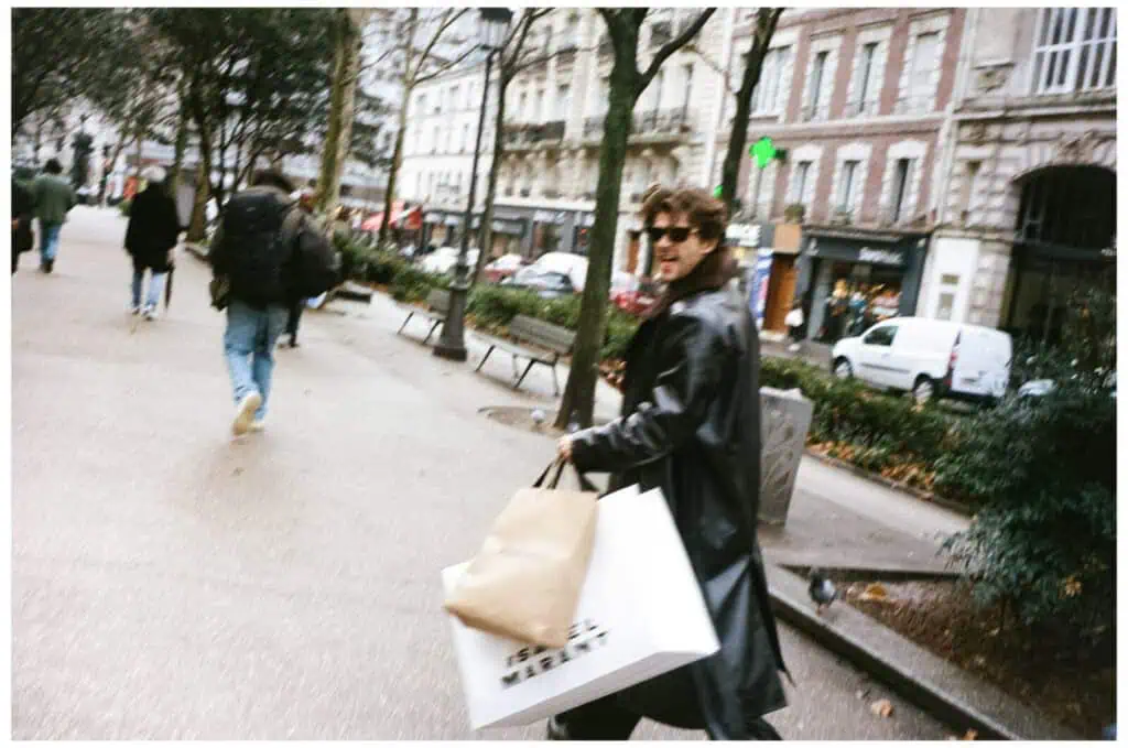 A person in a black leather coat and sunglasses walks down a city sidewalk, holding several shopping bags. Buildings, parked cars, and other pedestrians are visible in the background.