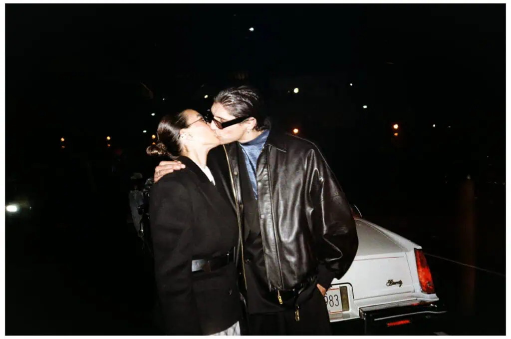 A couple kisses passionately at night in front of a white vintage car, illuminated by streetlights. Both wear dark clothing, and the man has sunglasses and a leather jacket.