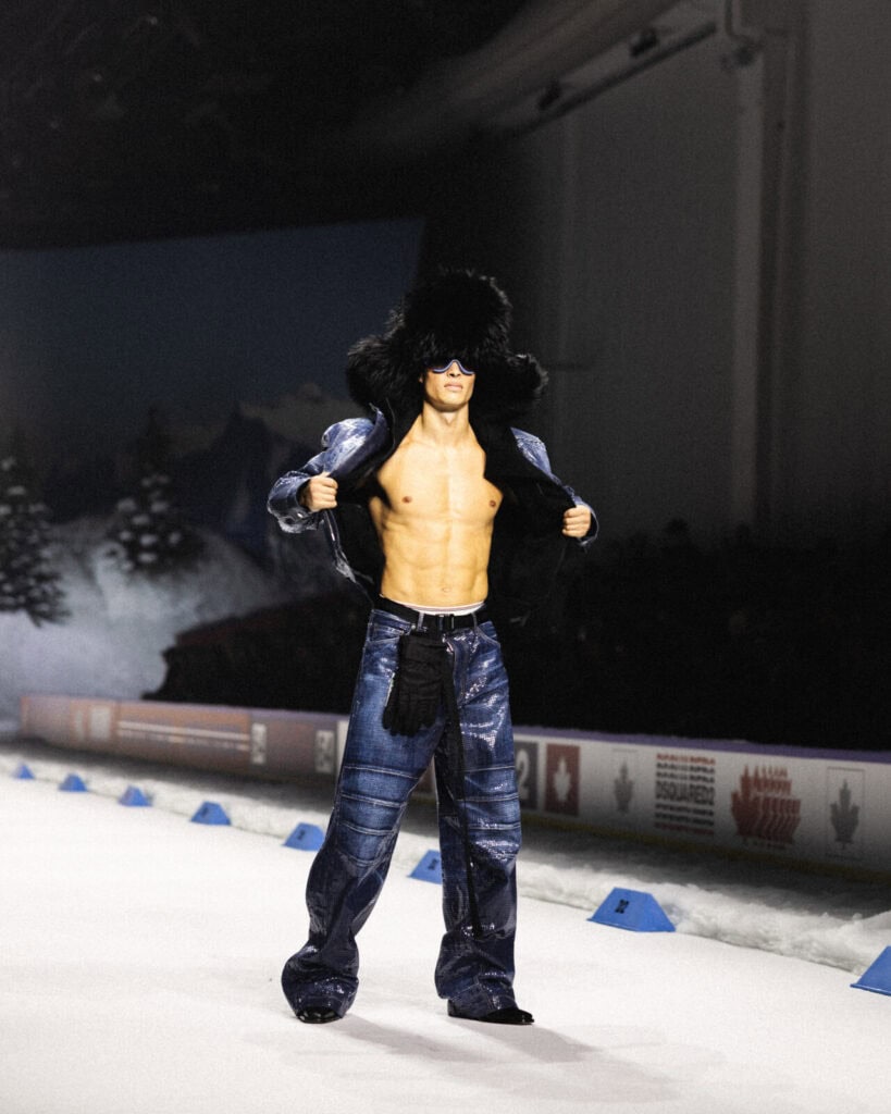 A shirtless male model on a runway wears shiny blue denim pants, an open matching jacket, large black boots, and a fluffy black hat, with a winter-themed backdrop and snow-covered trees behind him.