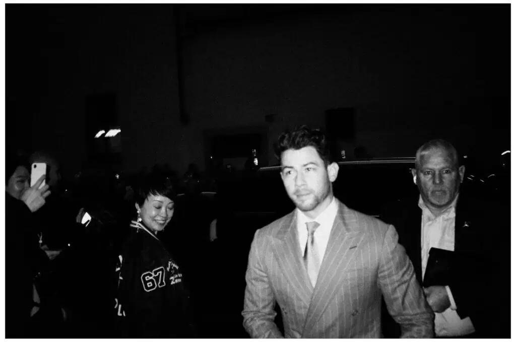 A man in a light pinstripe suit walks outside at night surrounded by people; some are smiling and taking photos. The scene appears crowded and lively.