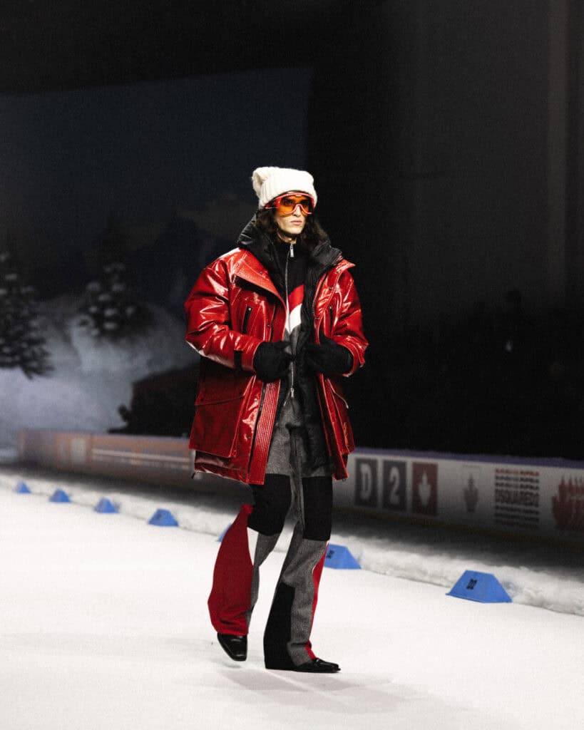 A model walks on a snowy runway wearing a shiny red puffer jacket, black layered outfit, white knit beanie, orange sunglasses, and heeled boots. Snowy trees and winter-themed decor are visible in the background.