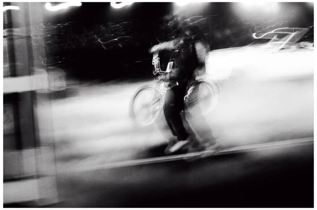 A blurred black-and-white photo showing a person walking with a bicycle at night, surrounded by bright lights and indistinct motion, creating a sense of speed and movement.