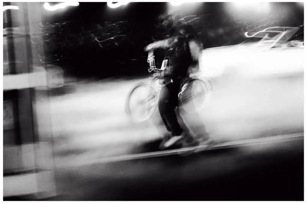A blurred black-and-white photo showing a person walking with a bicycle at night, surrounded by bright lights and indistinct motion, creating a sense of speed and movement.
