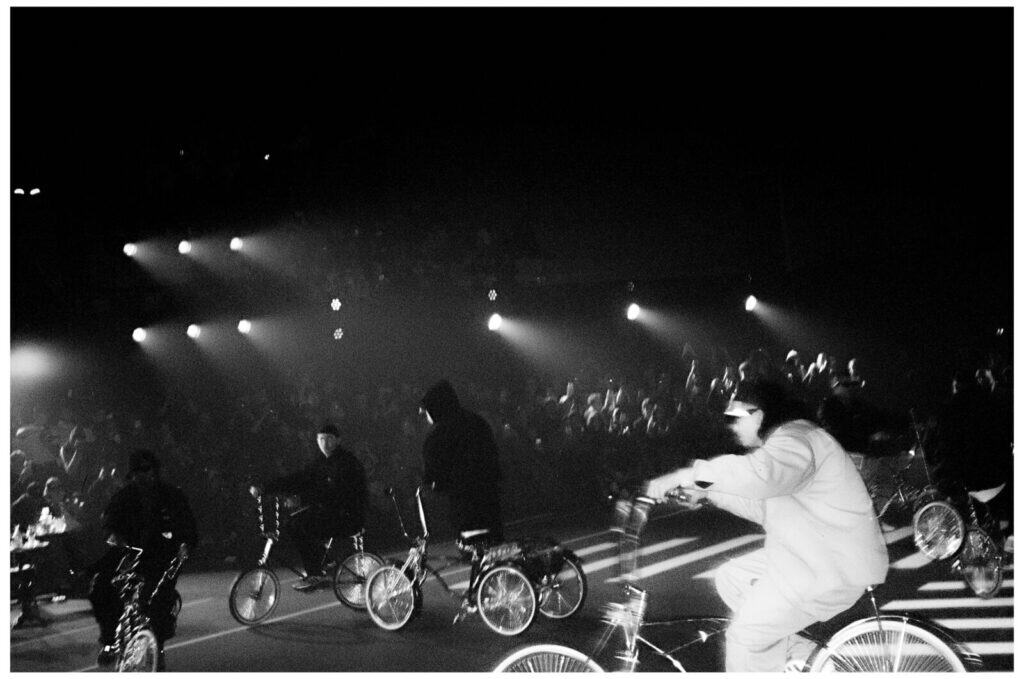 A black-and-white photo of several people riding bicycles indoors, with spotlights shining and a large crowd watching in the background. The scene appears lively and energetic, with dramatic lighting and a sense of motion.