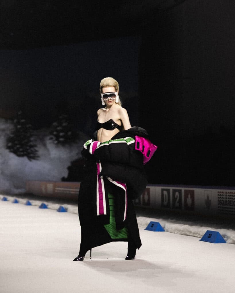 A model on a runway wears a dramatic black, pink, and green puffer coat over a black bra, with high-waisted pants, sunglasses, and tall, styled blond hair. The background features snow and dark, misty trees.