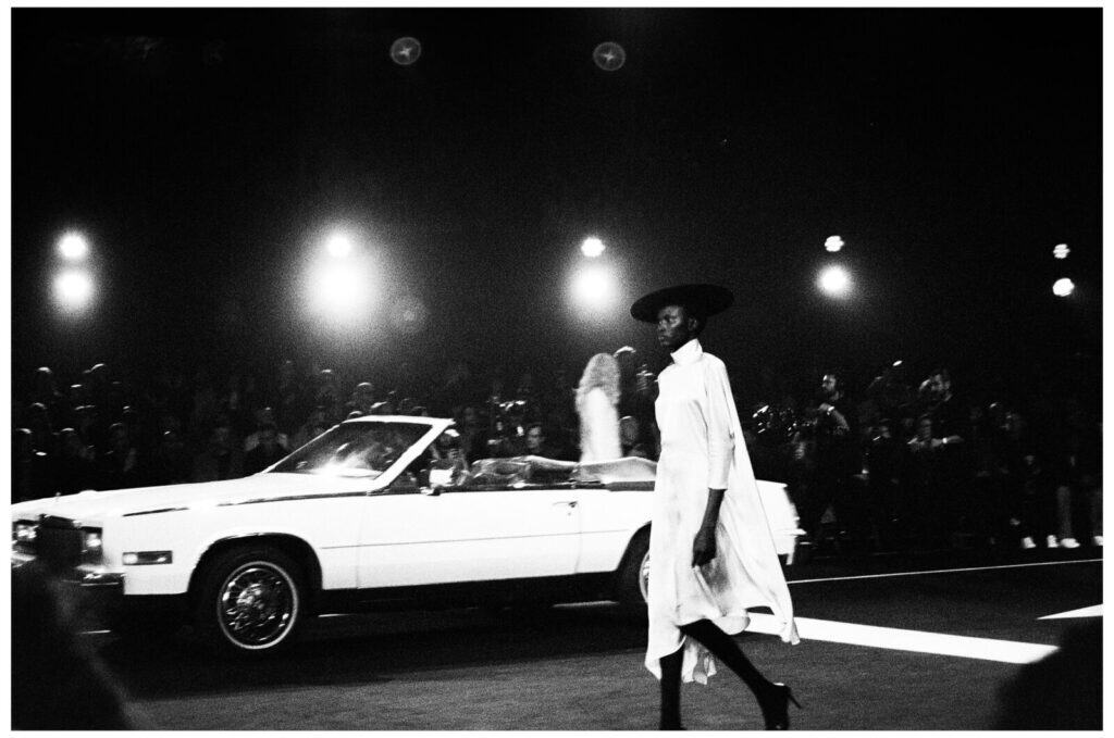 A model in a flowing dress and wide-brimmed hat walks on a runway in front of a white convertible car, with bright spotlights and an audience in the background. The image is in black and white.