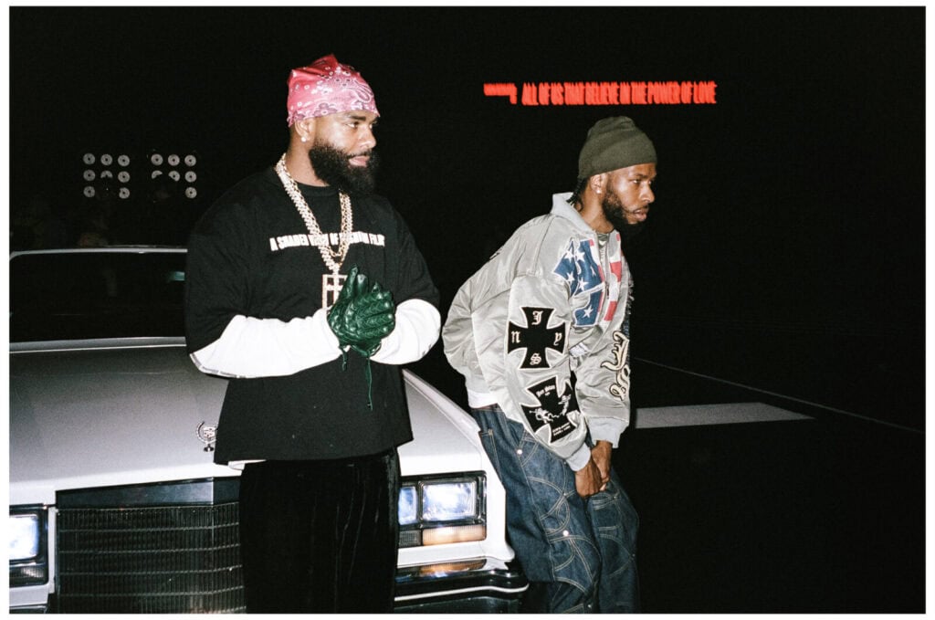 Two men stand in front of a white vintage car at night. One wears a pink bandana, black shirt, gloves, and jewelry; the other wears a gray hoodie with patches and a beanie. Red neon text glows in the background.