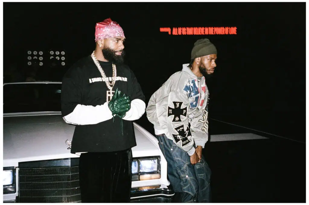 Two men stand in front of a white vintage car at night. One wears a pink bandana, black shirt, gloves, and jewelry; the other wears a gray hoodie with patches and a beanie. Red neon text glows in the background.
