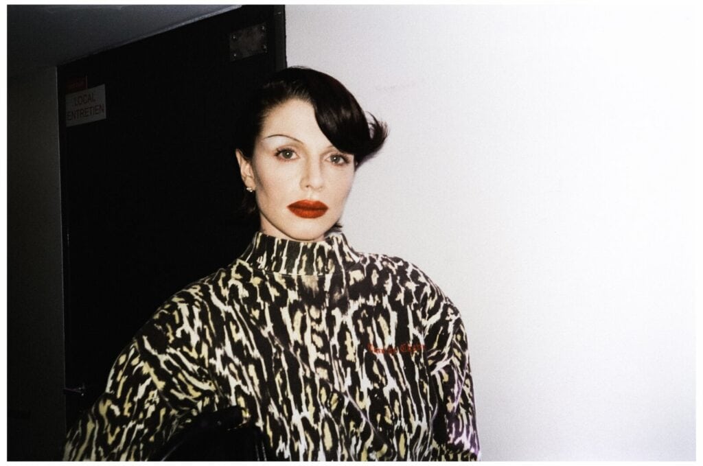 A person with short, dark, styled hair and bold red lipstick stands against a white wall, wearing a high-neck, animal-print top. The expression is neutral and the background features a partially visible dark door.