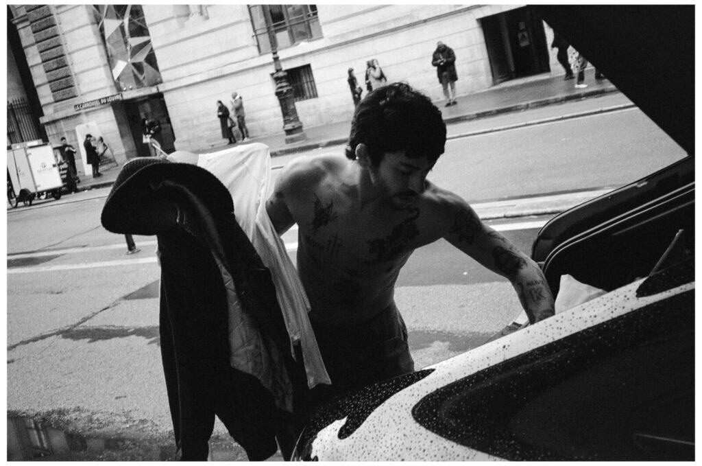 A shirtless man with tattoos stands by an open car trunk on a city street, holding clothing. Pedestrians and a building are visible in the background. The photo is in black and white.