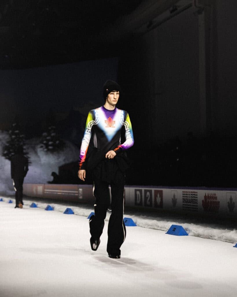 A person wearing a colorful, long-sleeved top with a red maple leaf design and black pants walks on an indoor ice rink, with snow-like decorations and winter scenery in the background.