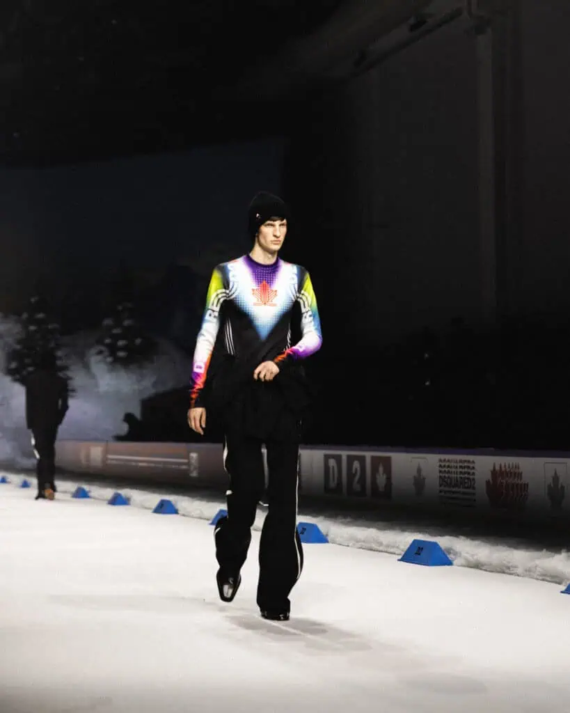 A person wearing a colorful, long-sleeved top with a red maple leaf design and black pants walks on an indoor ice rink, with snow-like decorations and winter scenery in the background.
