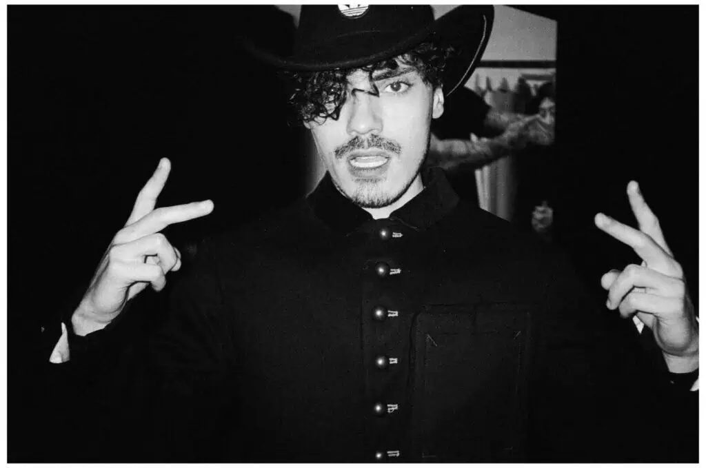 A person in a dark buttoned coat and wide-brimmed hat faces the camera, holding up both hands with peace signs. Their expression is intense, with slightly open mouth and curly hair partially covering one eye.